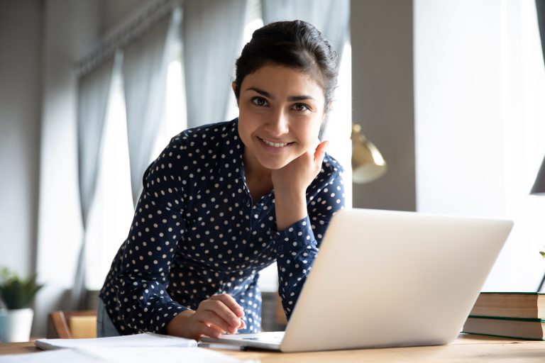 Portrait of smiling Indian girl standing at desk in living room working on laptop
